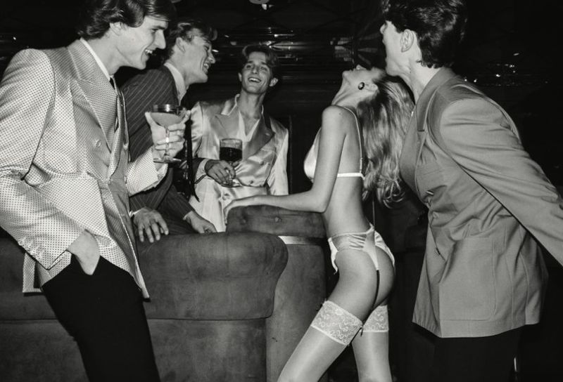 Monochrome image showing five adults gathered indoors at night; a woman in lingerie is leaning against a sofa, surrounded by suited men with drinks.