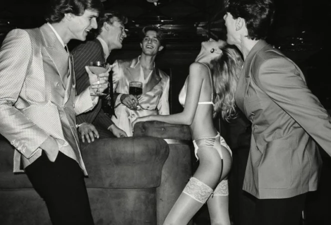 Monochrome image showing five adults gathered indoors at night; a woman in lingerie is leaning against a sofa, surrounded by suited men with drinks.