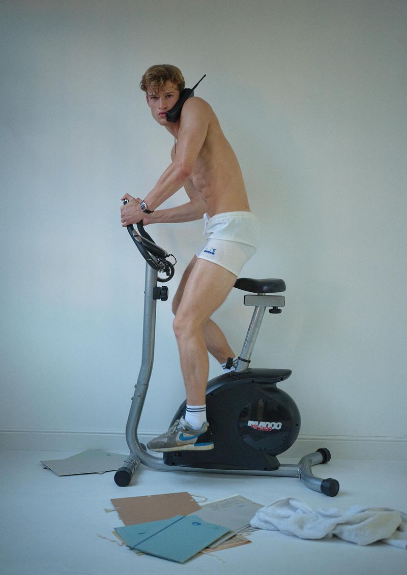 Athletic man on stationary bike with vintage phone. Shirtless adult male exercises indoors in white shorts, gray shoes, against white background.