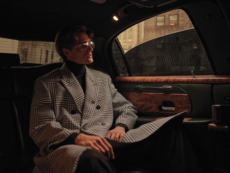 Man in patterned overcoat seated in a limousine looking out the window, styled with glasses and a turtleneck, shot in an urban setting.
