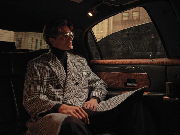 Man in patterned overcoat seated in a limousine looking out the window, styled with glasses and a turtleneck, shot in an urban setting.