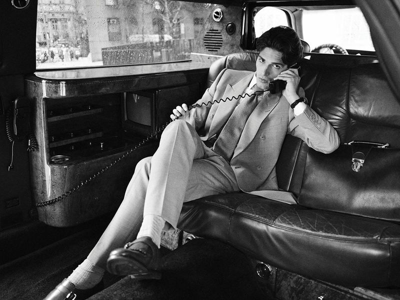 Monochrome photo of a man in a suit talking on the phone in the back of a limousine with city buildings seen through the window.