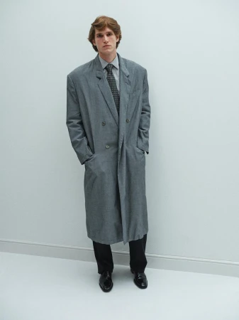 Model Alberto Paolini wearing an oversized grey overcoat, dark tie, and black trousers against a pale neutral background.