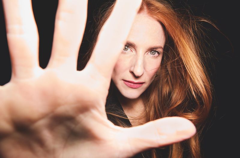 Portrait of red-haired woman with her palm near the camera. Red hair is framing her face. She has fair skin, a black background.