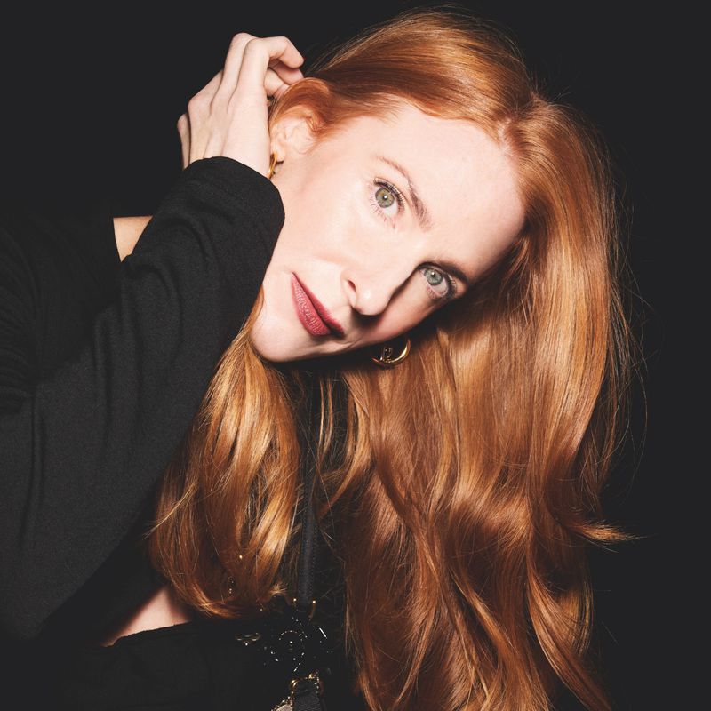 Headshot of a woman with long, red wavy hair looking at the camera with a dark background, wearing a black top and gold earrings.