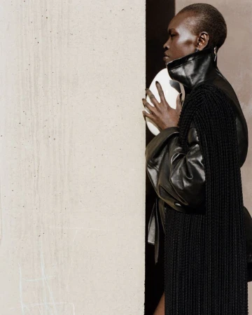 Fashion shot of black female adult holding white spherical prop, wearing black leather-look top and fringed shawl in studio.
