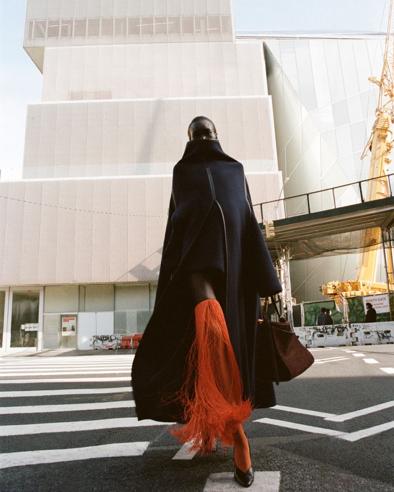 Woman in dark coat and orange fringed skirt walks across crosswalk in front of modern architecture with a construction crane nearby.