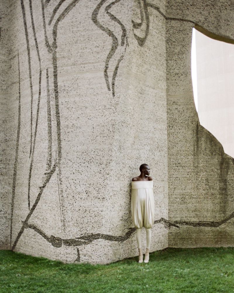 A model in an off-white dress and shoes poses outdoors against a textured concrete wall with a drawing and green grass.