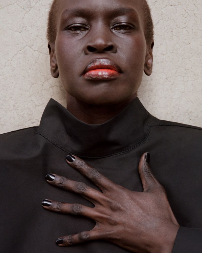 Close-up portrait of a dark-skinned adult female with short hair, wearing a black turtleneck, and her hand resting on her chest.