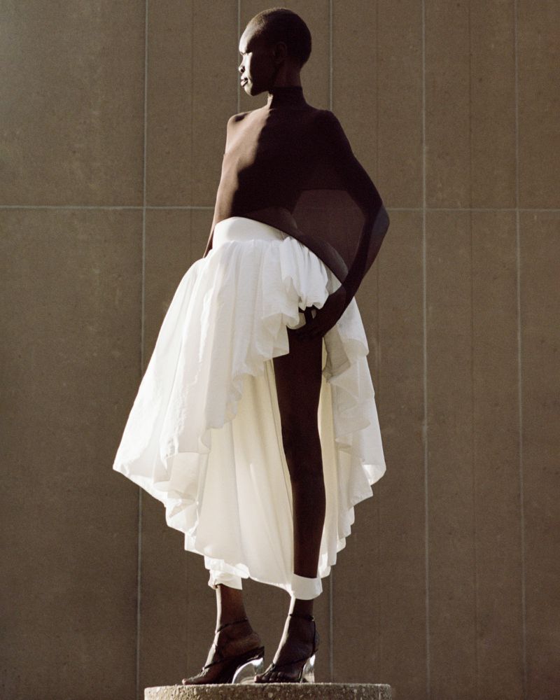 Fashionable African American woman in a brown bodysuit and layered white skirt posing outdoors on a concrete platform in sunlight.
