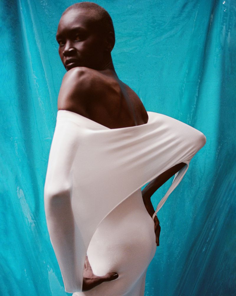 African American woman poses in a studio wearing an off-the-shoulder white dress against a blue textured background.