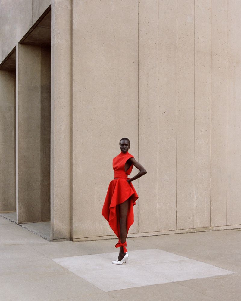 Fashion shoot of a slender Black woman in a red dress with a high neckline and asymmetrical hem standing on a concrete floor.