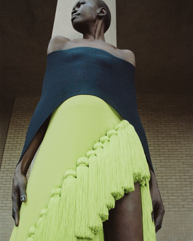 Fashion shot of a Black woman in a green and black dress with tassels, standing against a neutral brick wall outdoors in daylight.
