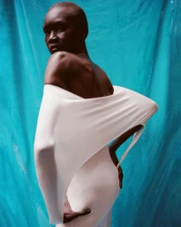 Model Alek Wek wearing a sculptural off-the-shoulder white dress against a vibrant draped cyan fabric backdrop.
