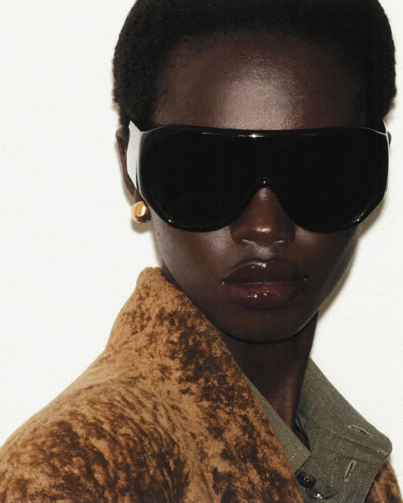Close-up studio shot of a dark-skinned woman wearing oversized black sunglasses, gold earrings, and a brown textured coat against a white background.