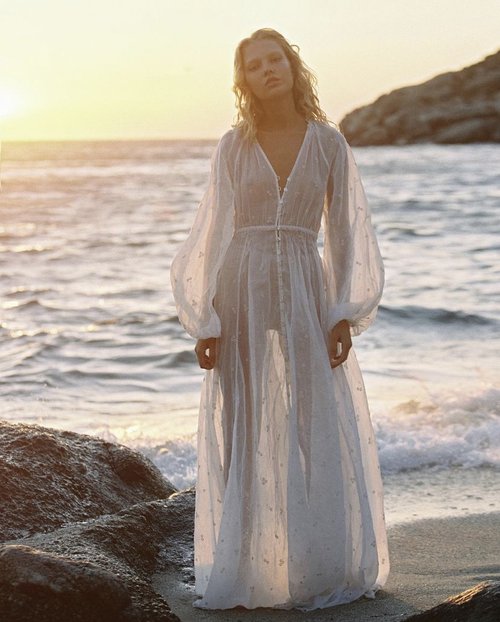 Woman in a sheer white dress stands on a beach at sunset with waves, rocks, and distant land visible in the background, golden light.
