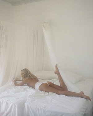 Back view of a woman lying on white linens in a bedroom with a white curtain canopy and white walls, indoors.