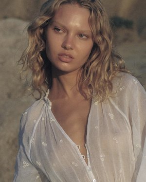 Close-up portrait of a woman with wavy blonde hair wearing a sheer white embroidered blouse outdoors in natural sunlight.