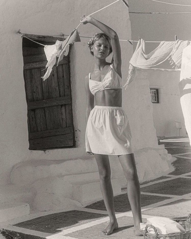 A woman stands outdoors in a two-piece bathing suit next to a building with a clothesline and hanging laundry in a fashion shot.