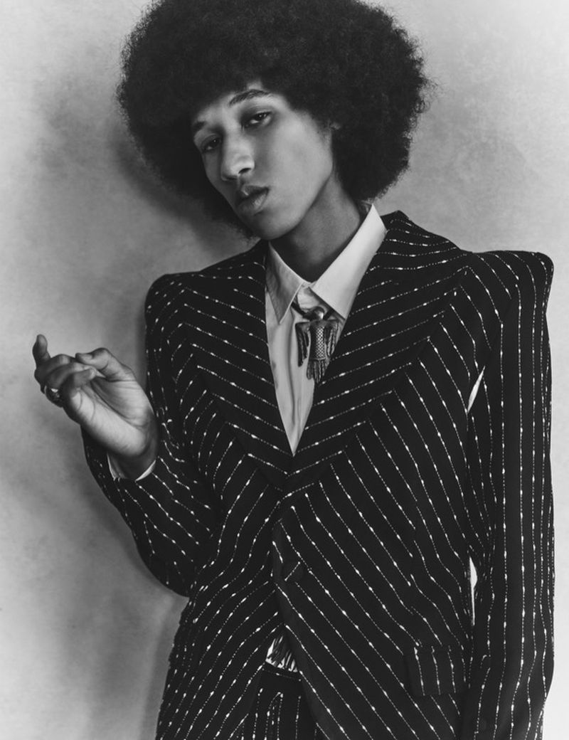 Monochrome portrait of an adult with large afro wearing a striped suit and bolo tie against a textured grey wall.