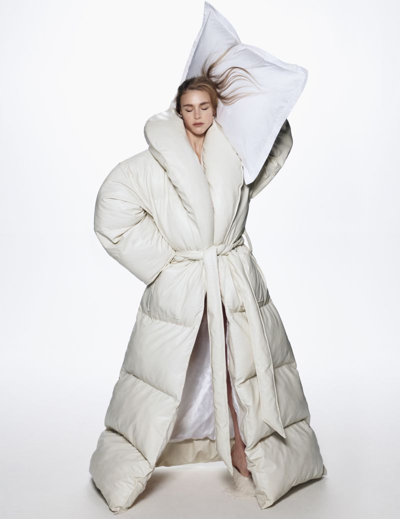 Woman in a puffy white robe poses with a pillow against a white background, creating a surreal fashion portrait.