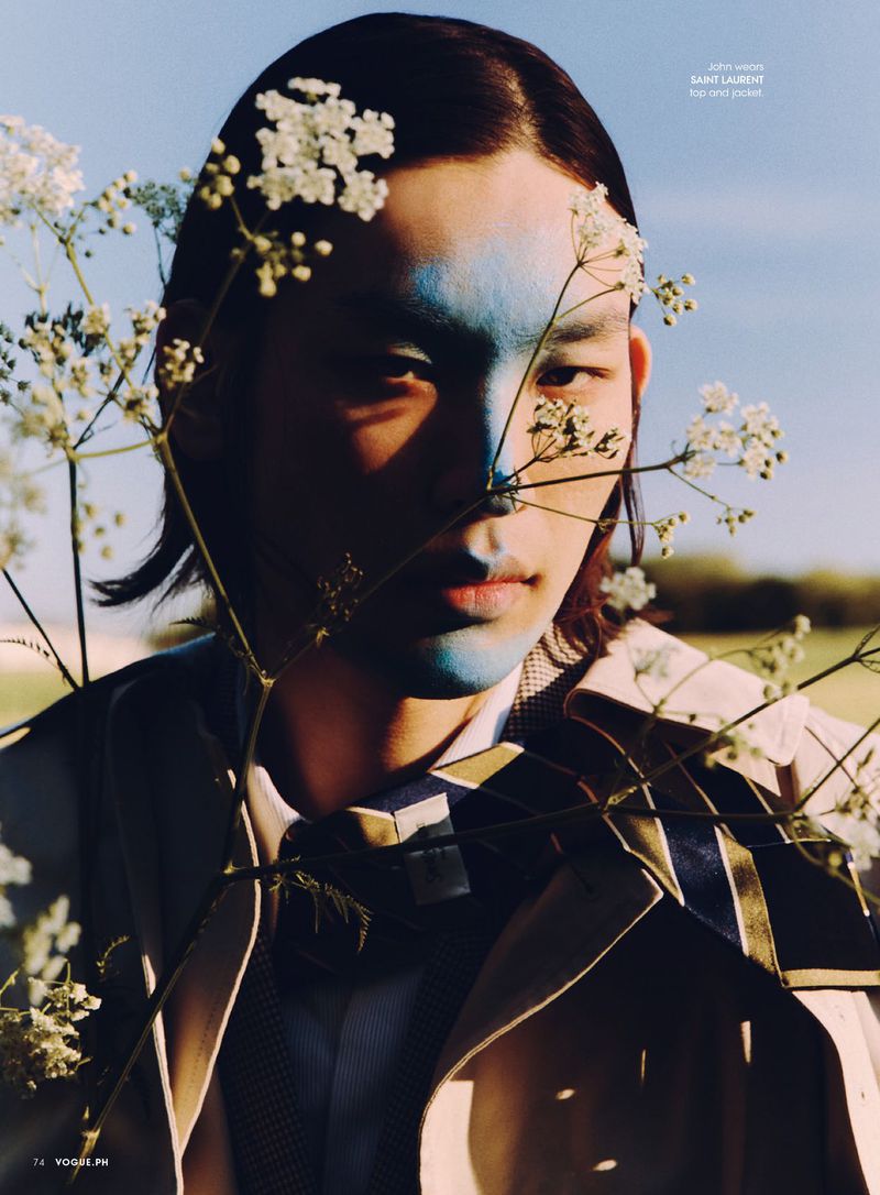 Model John Ganbat with blue facial pigment looking through white wildflowers wearing a beige trench coat for Vogue Philippines.