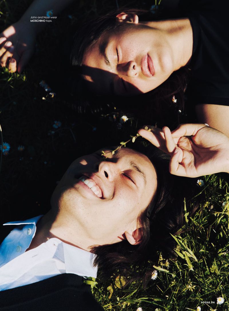 Two young adults lie on a sunlit meadow, wearing stylish outfits, captured in a close-up shot with a shallow depth of field. 