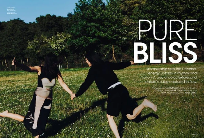 Two women in dresses holding hands and running through a grassy field with trees in the background, fashion editorial, outdoor setting.