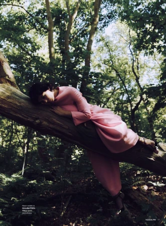 Fashion editorial shot of a woman in a pink dress resting on a fallen tree trunk in a sun-dappled forest setting.