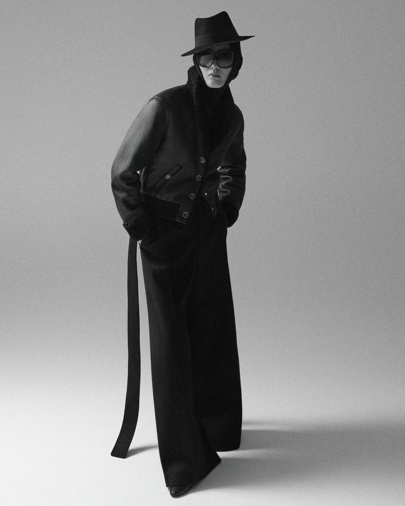 Monochromatic fashion shot of a model in black hat, sunglasses, jacket, and wide-leg trousers against a neutral gray background in the photo studio.