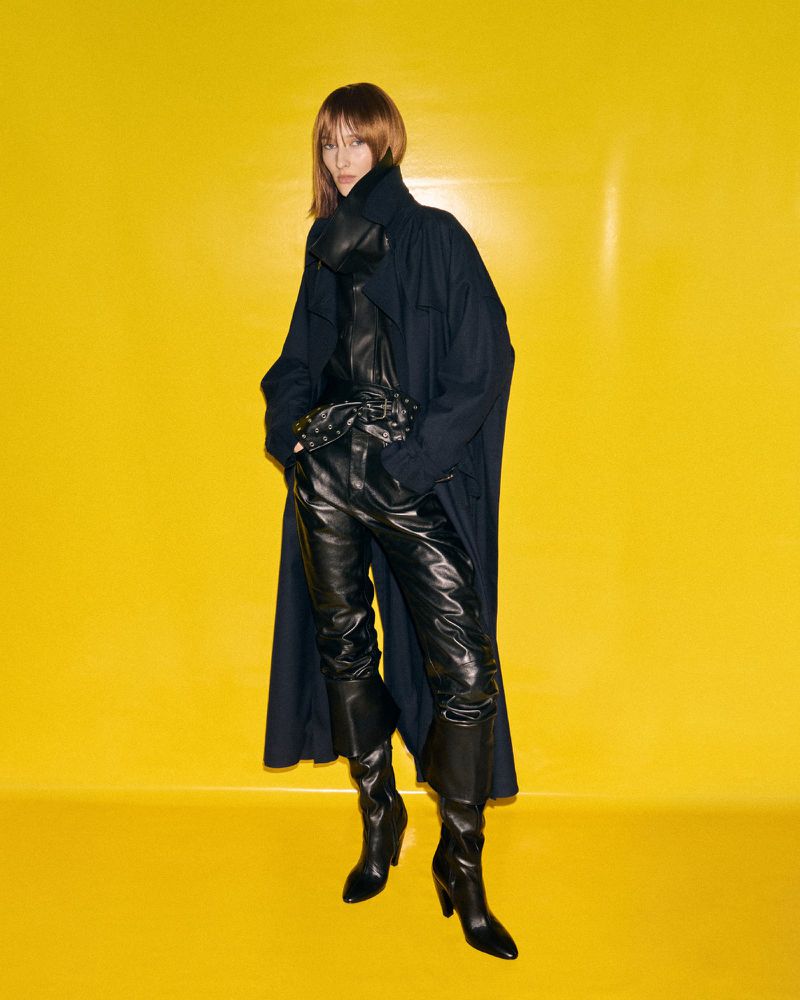 Stylish adult woman in a long dark coat, leather pants, and high-heeled boots strikes a pose against a vibrant yellow backdrop indoors.