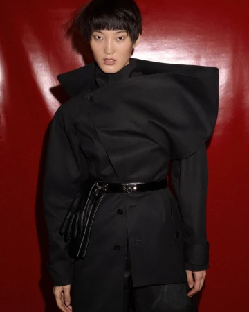 Model Wang Fei posing in a tailored black sculptural coat and glossy belt against a deep red background.
