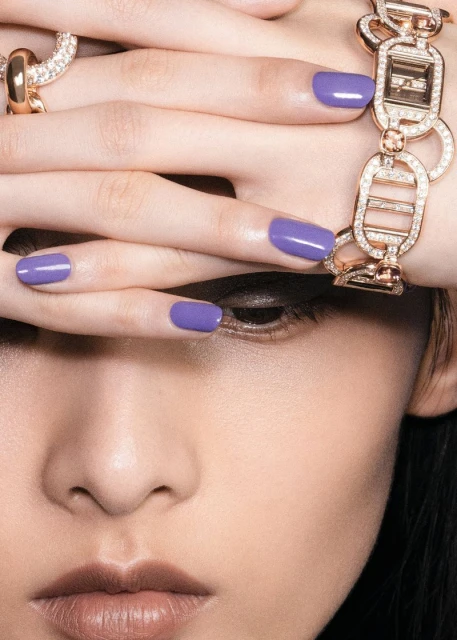 Close-up of a woman's face with purple nails, gold bracelet, and ring in a beauty or fashion editorial style image.