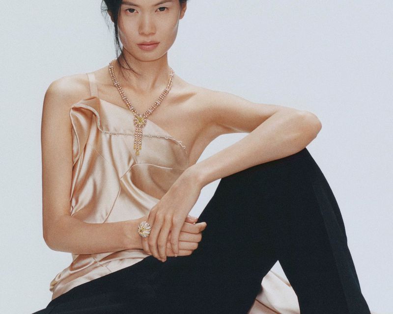 Elegant Asian woman poses wearing a gold necklace and ring, a flowing asymmetrical peach top, and black pants against a white backdrop.