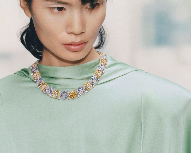 Close-up of a woman wearing a colorful floral necklace and a mint-green dress, highlighted by soft lighting in a bright, neutral setting.