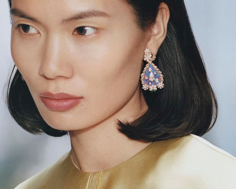 Portrait of an Asian woman wearing ornate, floral earrings, fair complexion, gold dress, looking to the side against a light blue background.