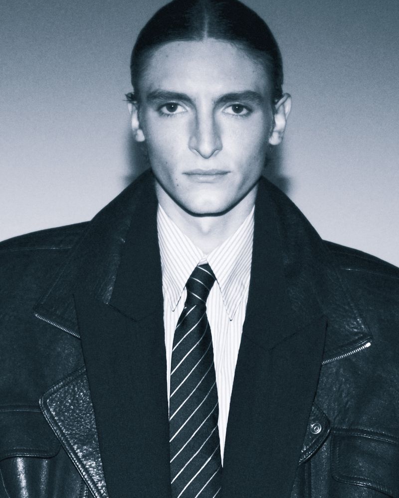 Portrait of an adult man in a striped dress shirt, suit jacket, and leather motorcycle jacket, looking at the camera with a neutral expression.