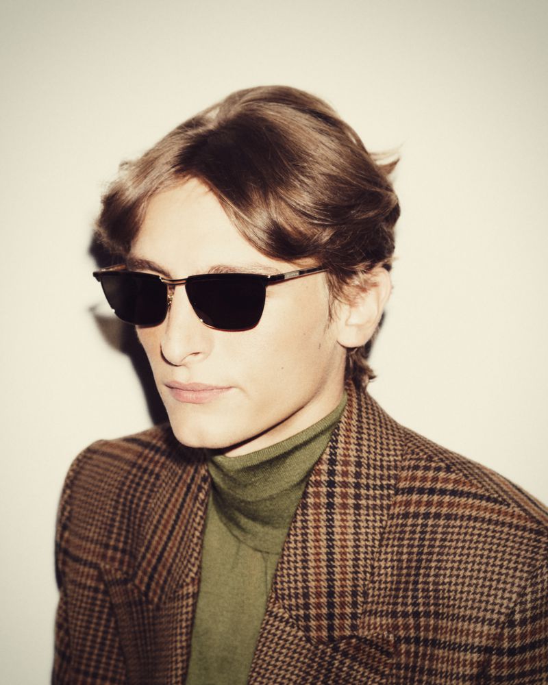 Person in sunglasses and plaid jacket poses against a white backdrop, capturing a stylish, retro-inspired fashion moment.