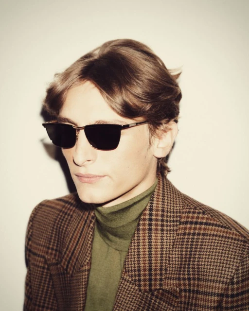 Person in sunglasses and plaid jacket poses against a white backdrop, capturing a stylish, retro-inspired fashion moment.