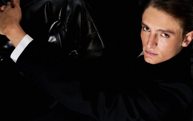 Portrait of a fair-skinned adult male in a black turtleneck and jacket, posing in a dark indoor setting with subdued studio lighting.