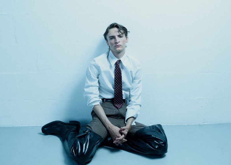 A young adult man is sitting on the floor with his knees up, wearing a striped shirt, tie, and large black boots against a plain background.