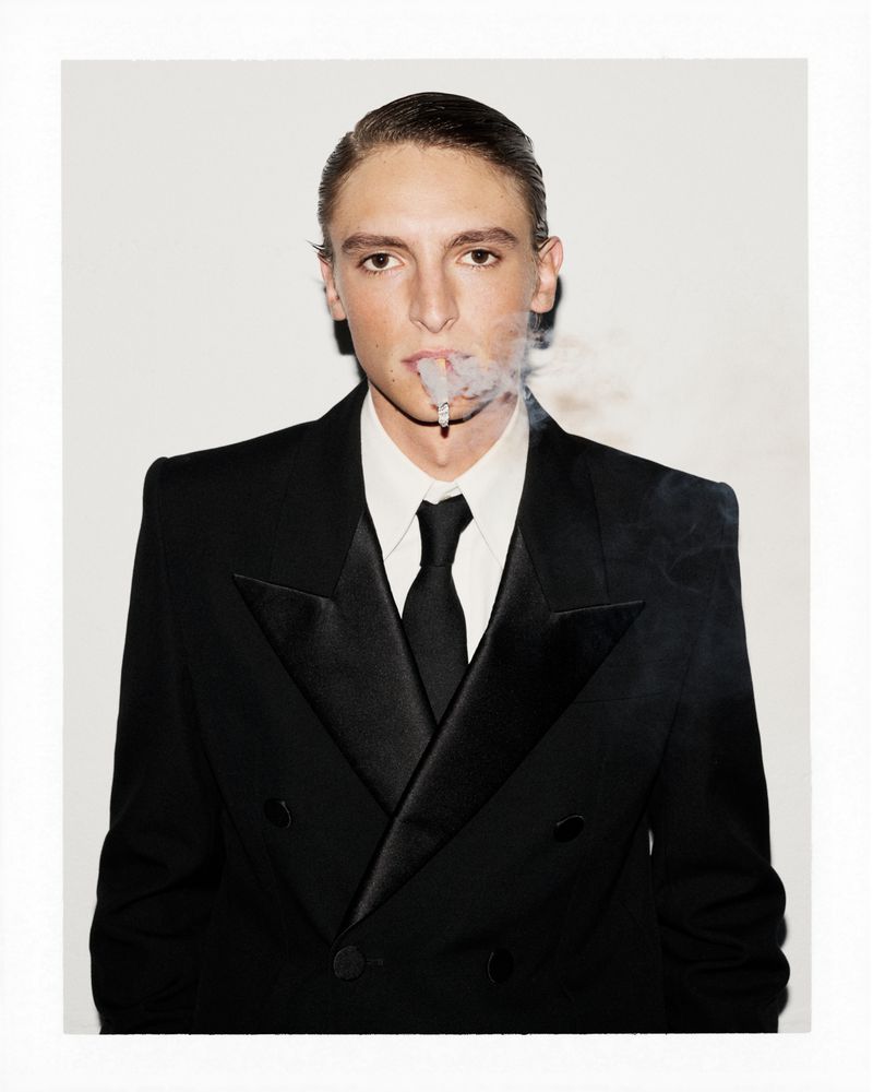 Portrait of a stylish young man smoking a cigarette in a black tuxedo and tie, against a bright white background in a studio shot.