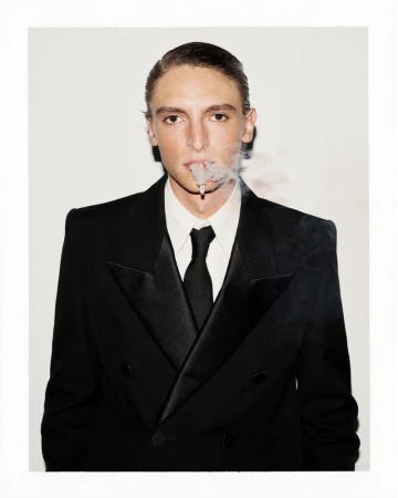 Portrait of a stylish young man smoking a cigarette in a black tuxedo and tie, against a bright white background in a studio shot.