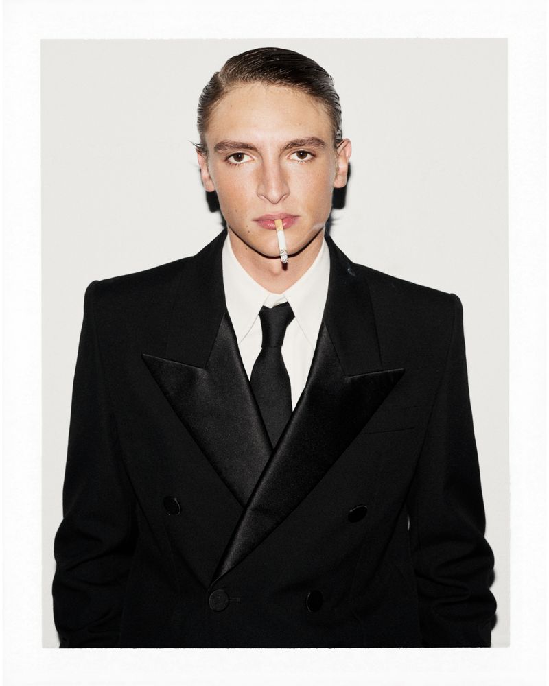 Portrait of a young adult male in a black suit with a cigarette in his mouth against a white background in a photo studio.