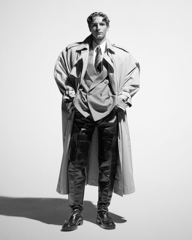 Black and white studio shot of a male model wearing a tan trench coat, grey suit, black pants, and boots, hands in pockets.