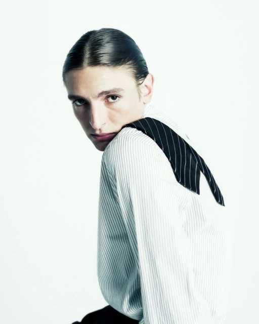 Portrait of a young adult with slicked-back hair wearing a vertically striped shirt, posing in a bright, neutral studio environment.