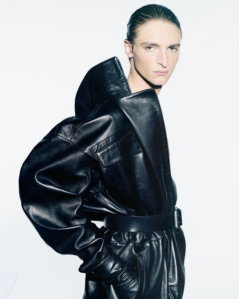 Model poses in a black leather jacket and pants against a bright white background, showcasing fashion and style with a sleek and modern aesthetic.