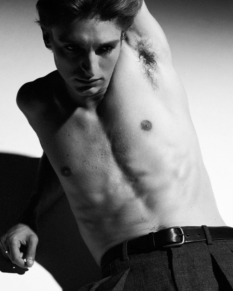 Black and white portrait of a shirtless adult male model with dark hair posing indoors, wearing pants and a belt, and set against a white background.