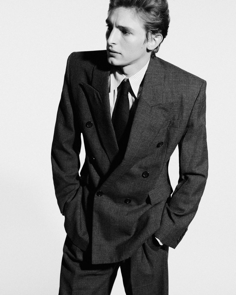 Monochrome portrait of a young adult male with wavy hair, wearing a double-breasted suit with hands in pockets, against a white backdrop.