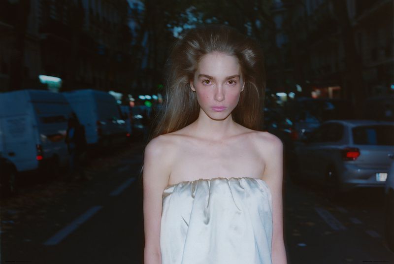 Portrait of a fair-skinned woman with long brown hair, wearing a draped white top, standing on a city street at night. Cars are in the background.